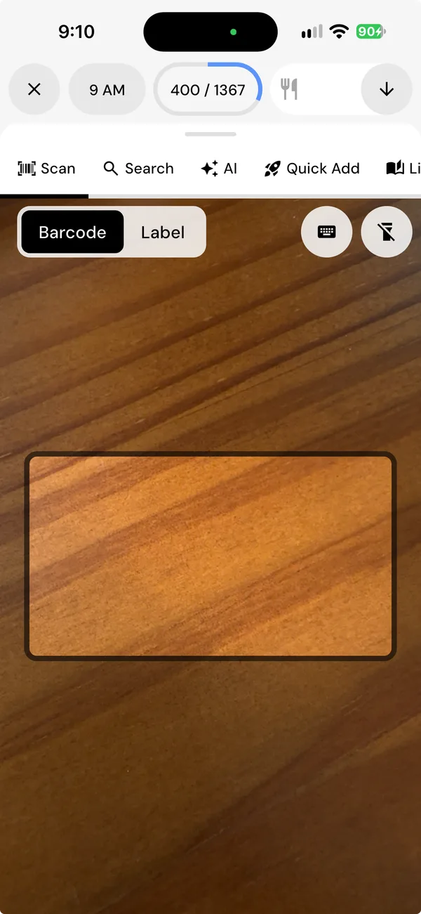 MacroFactor's Scan tab on Barcode, with a Label option beside it, and the camera framing a box over a wooden table.